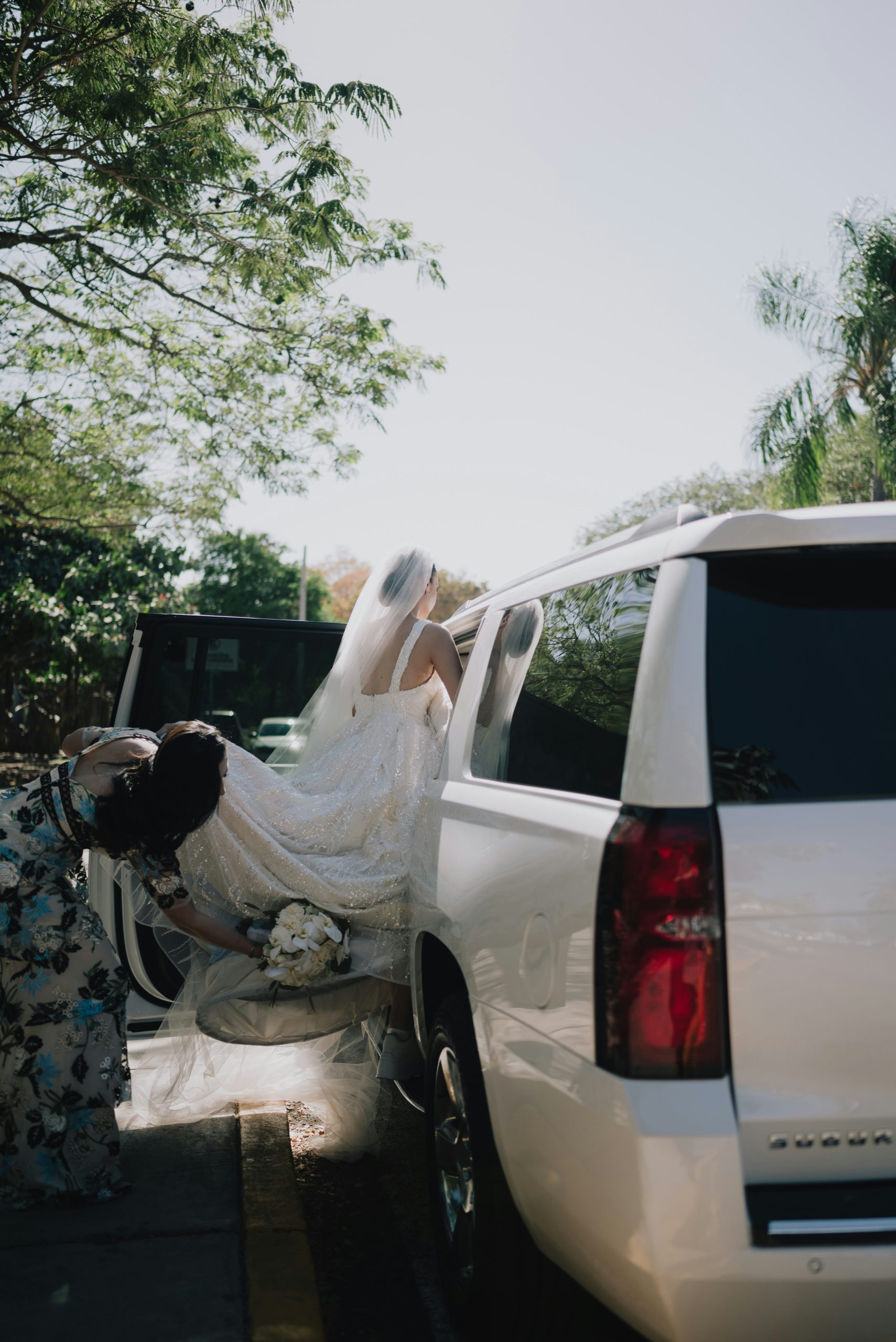 A bride in a shimmering white wedding gown with a long train being assisted into a white luxury SUV for her wedding and special event transportation.