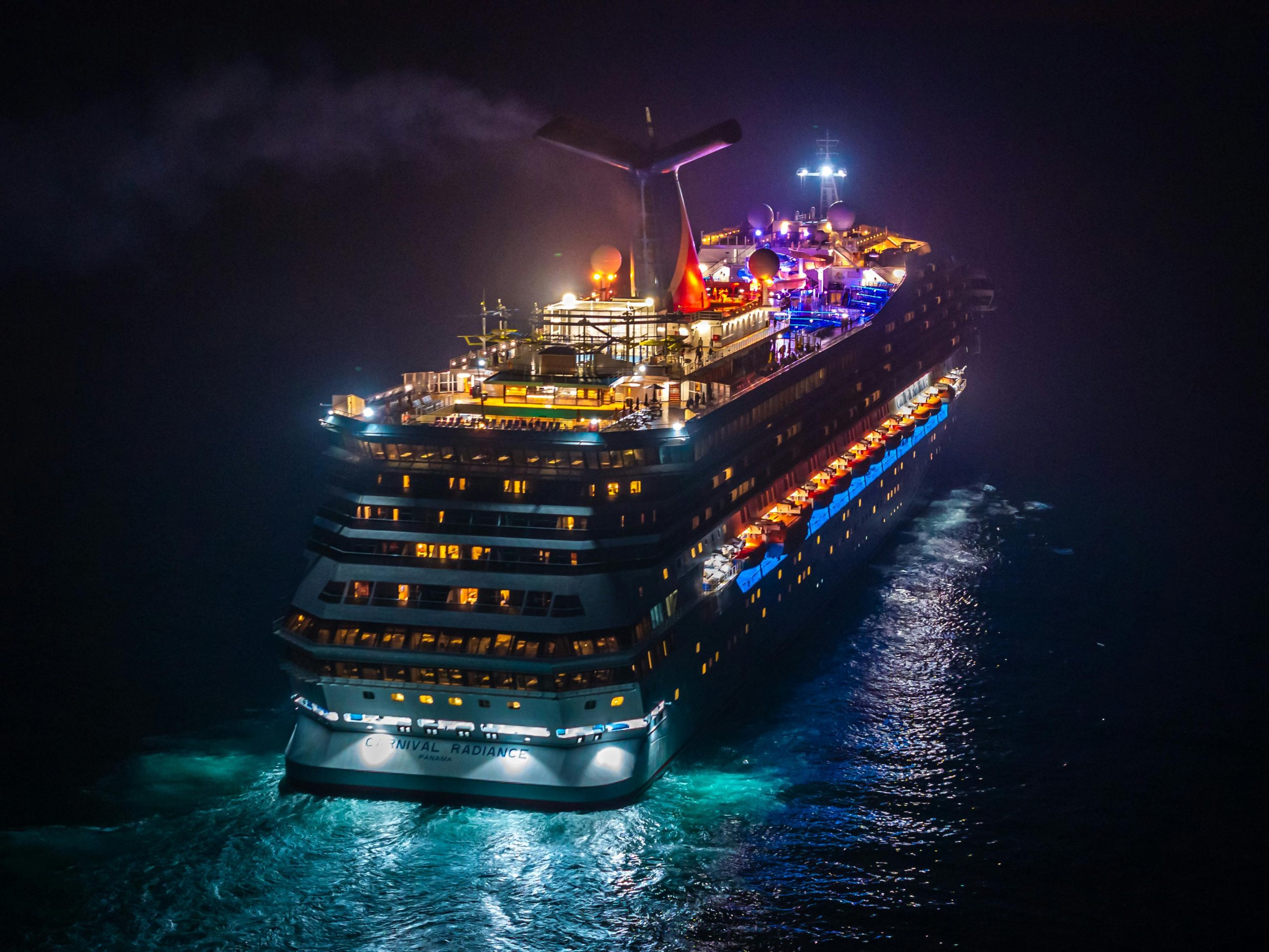 An aerial night view of the illuminated Carnival Radiance cruise ship sailing on dark water, representing Orlux's professional port transfer services.