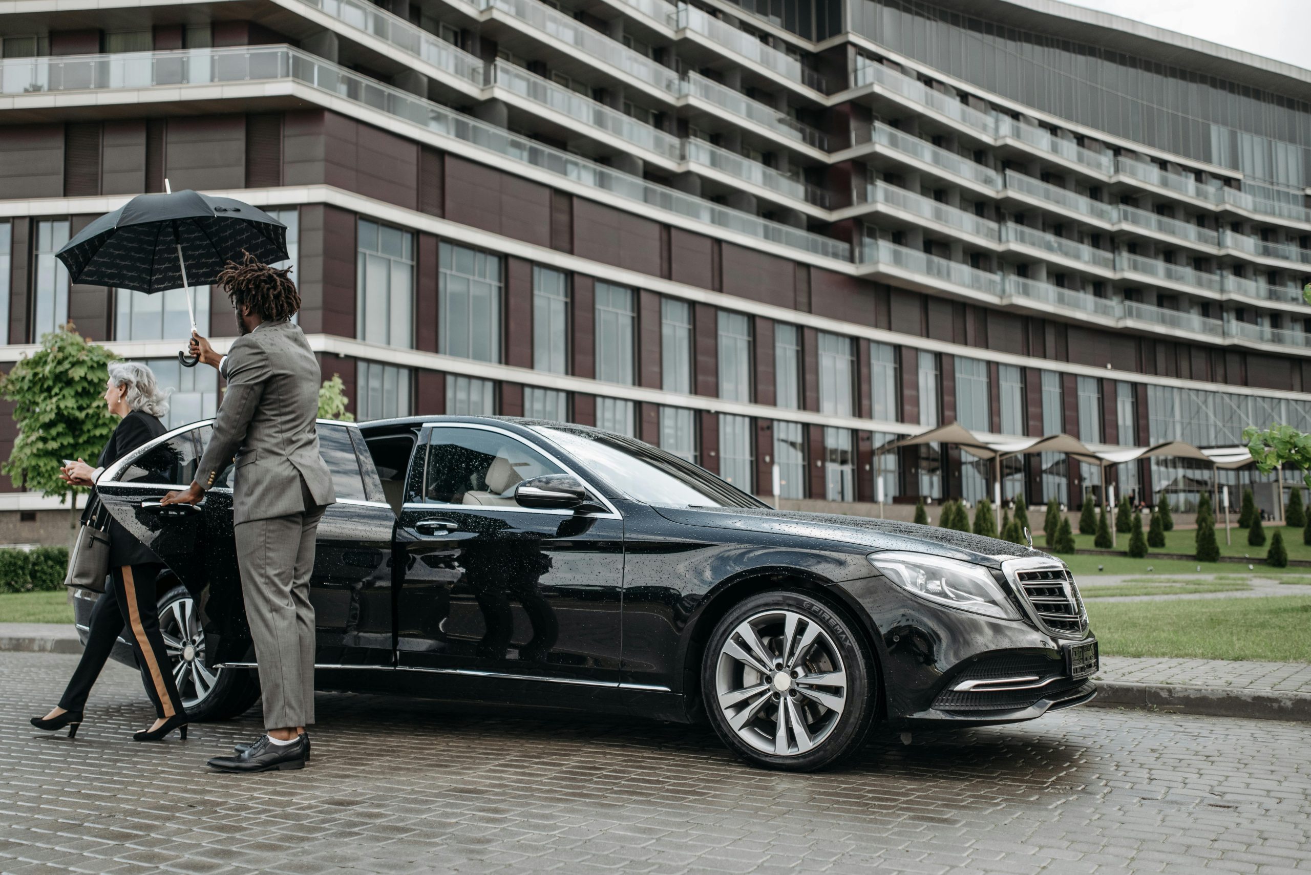 A professional Orlux chauffeur in a grey suit holds a black umbrella for an elderly female passenger getting into the back seat of a black luxury sedan outside a modern hotel.