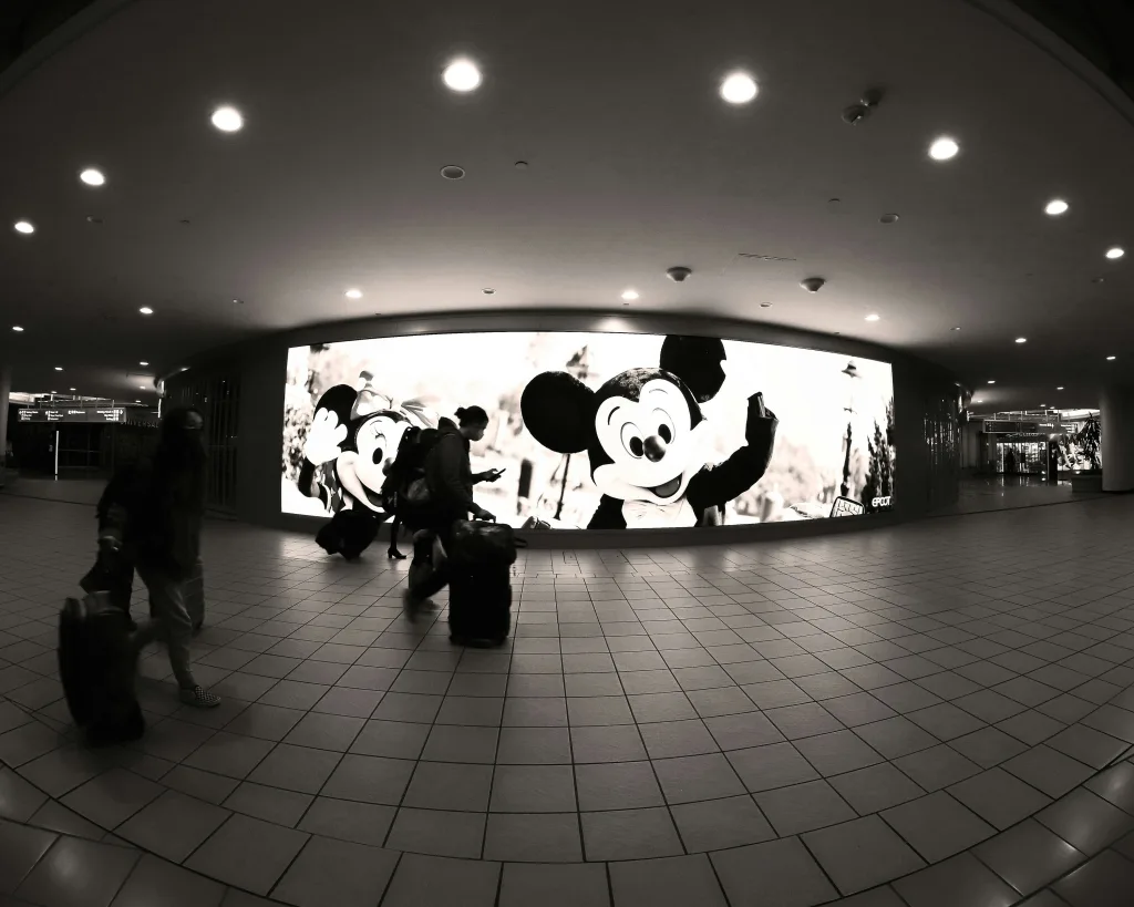 Travelers with luggage in an airport terminal, passing by a large display featuring Mickey Mouse, promoting a family-friendly atmosphere for Orlando Airport (MCO) visitors.