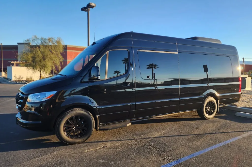 Black high-roof Mercedes-Benz Sprinter passenger van with black wheels, parked in an open lot at sunset